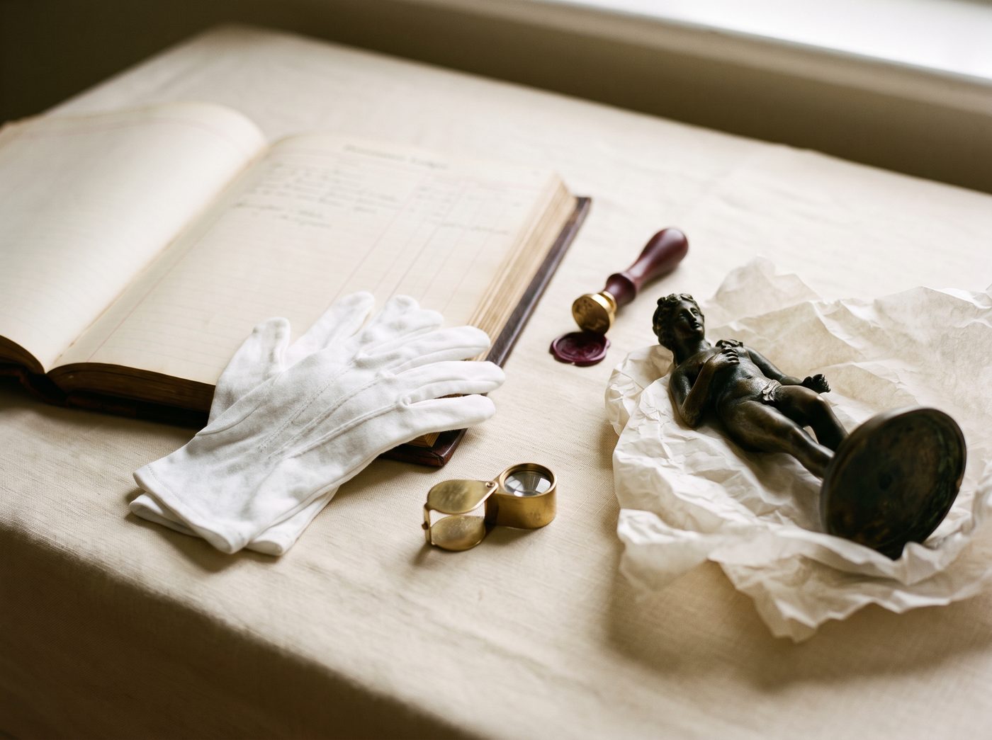 White archival gloves, a provenance ledger, a wax seal and a small bronze sculpture on a linen-covered table